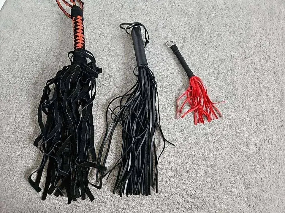 Flogger-style tools on a carpet, including two black fringe floggers and a smaller red tassel whip